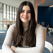 Camryn Amens sits in the lobby of the Health Sciences Building, smiling to the camera 